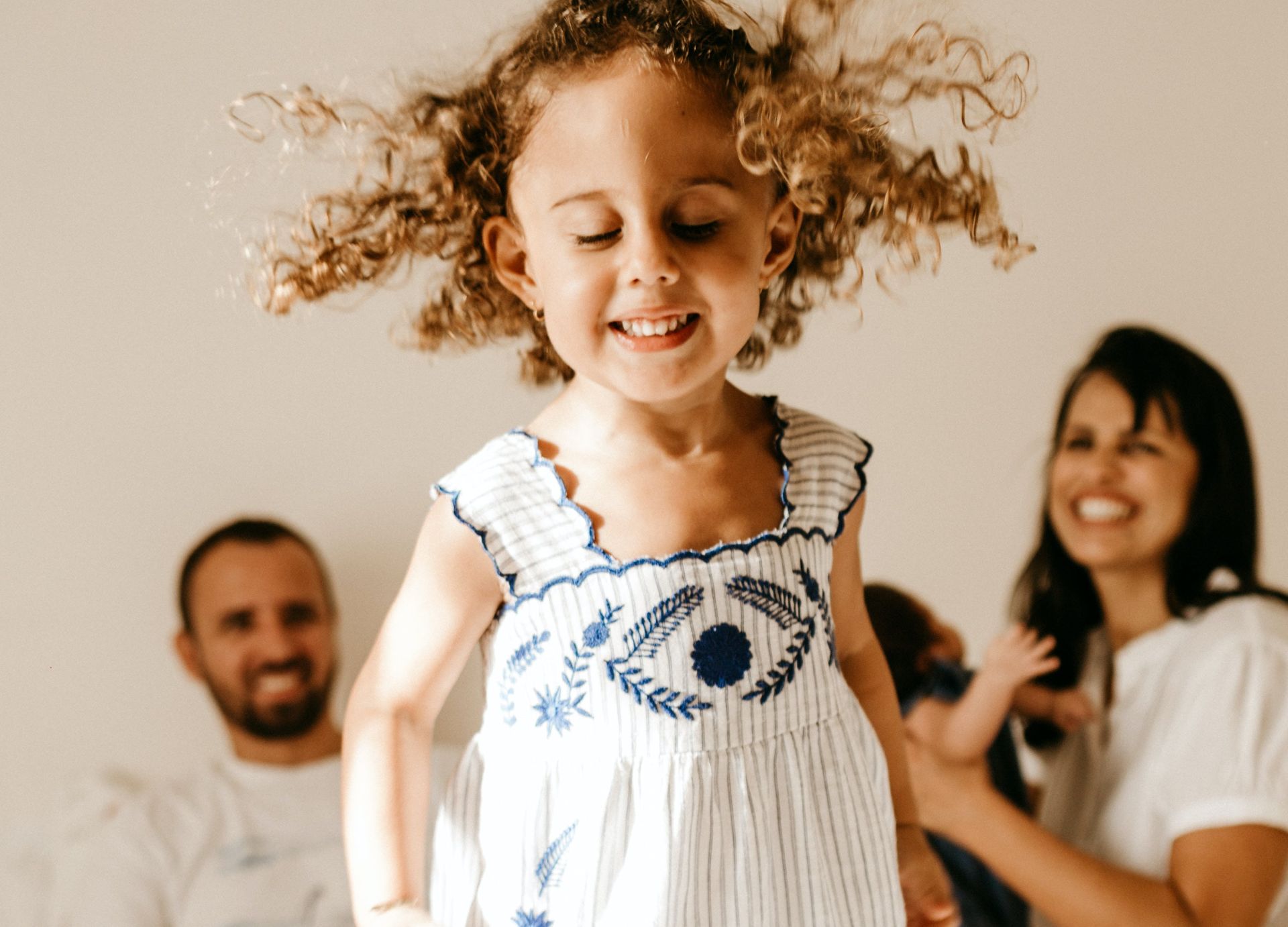 Adorable little girl with curly hair in light dress jumping on bed with closed eyes near happy young parents and newborn baby